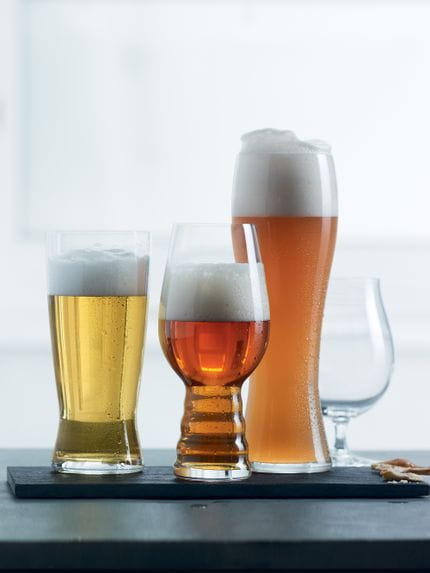 Vier SPIEGELAU Beer Classics Gläser in einer Reihe eins ist leer und der Rest mit verschiedenene Bieren gefüllt. Die drei gefüllten Gläser stehen auf einer schwarzen Servierplatte vor einem hellen Hintergrund.<br/>