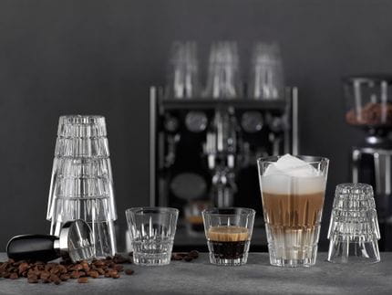 Le verre à espresso de la collection SPIEGELAU Perfect Serve rempli d'espresso et le verre Highball rempli de Latte Macchiato sur un comptoir de bar décoré de grains de café et d'un tamper. En arrière-plan, une machine à café et un moulin.<br/>
