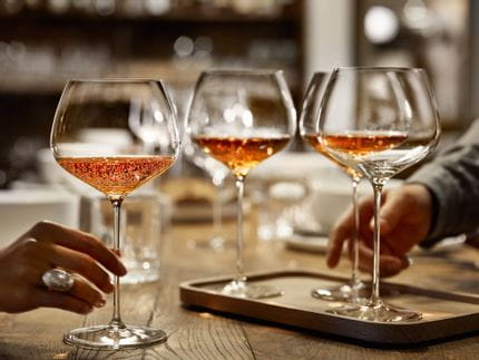 Una mano sostiene la copa de Borgoña SPIEGELAU de la serie Willsberger Anniversary, llena de vino espumoso de color rosado. Más atrás, en la mesa de madera, hay más copas.<br/>