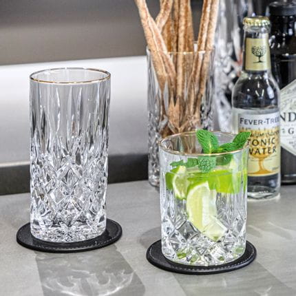 Das NACHTMANN Noblesse Gold Longdrinkglas neben dem mit einem Gin-Tonic-Cocktail gefüllten Trinkglas. Im Hintergrund ein weiteres Longdrinkglas, gefüllt mit Crissini und einer Flasche Tonic Water.<br/>
