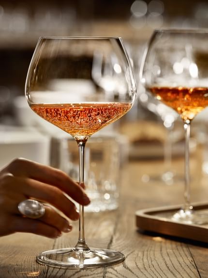 A hand holding the SPIEGELAU Burgundy glass of the series Willsberger Anniversary, filled with rosé colored sparkling wine. Further back on the wooden table, there are more glasses.<br/>