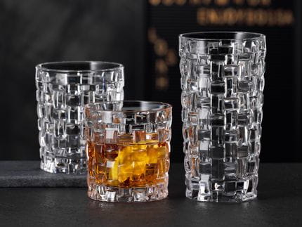 Il NACHTMANN Bossa Nova SOF riempito di whisky accanto al bicchiere longdrink Bossa Nova vuoto su una credenza in pietra nera. Dietro di loro il tumbler Bossa Nova vuoto.<br/>