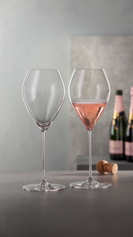Two SPIEGELAU Spumante glasses standing on a table with a cork in the background. One of the glasses is filled with a pink sparkling wine.<br/>