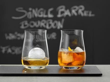 Two SPIEGELAU stemless Bourbon glasses, filled with whiskey and ice cubes. In the background a blackboard with Single Barrel Bourbon written on it.<br/>