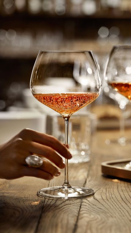 Una mano tiene il bicchiere SPIEGELAU Burgundy della serie Willsberger Anniversary, riempito di spumante rosé. Più indietro, sul tavolo di legno, ci sono altri bicchieri.<br/>