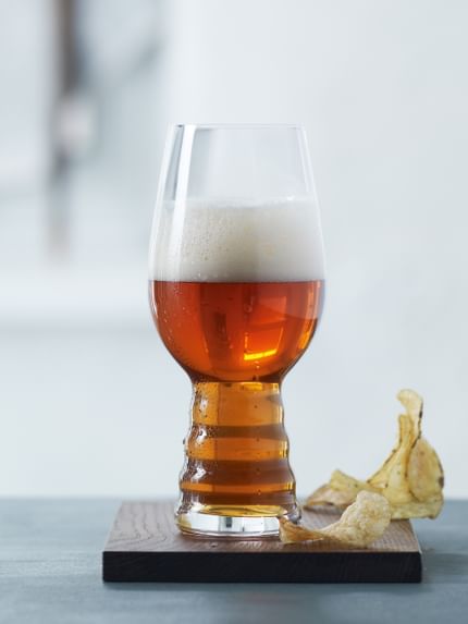 Un grupo de vasos de cerveza artesanal SPIEGELAU para IPA sobre una mesa. Uno de pie sobre un posavasos de madera, lleno de cerveza IPA. Junto a este vaso hay unas patatas fritas.<br/>