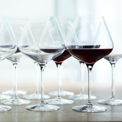 A group of SPIEGELAU Hybrid Burgundy glasses on a wooden table. Some of the glasses are filled with red wine, some are empty.<br/>