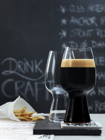 Two SPIEGELAU Craft Beer Glasses for Stout on a slate tray. One of them is filled with dark Stout beer, the glass behind is empty. Behind them is a small bag with potato chips and a blackboard with a beer menu on it.<br/>