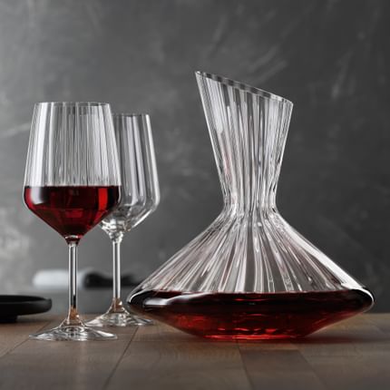 The with red wine filled SPIEGELAU Lifestyle decanter on a table. Left of the decanter two Lifestyle red wine glasses, one of them filled with red wine. On the left edge of the table an empty black plate.<br/>