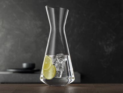 Il sottile decanter SPIEGELAU Style riempito con acqua, fette di limone e cubetti di ghiaccio su un tavolo di legno.<br/>