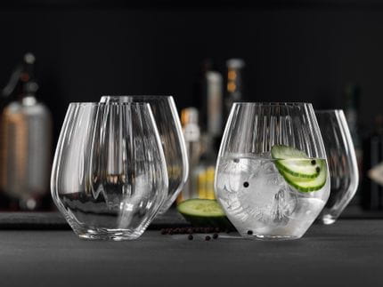 Four SPIEGELAU Gin and Tonic tumblers with optical line effects in the glass design on a table. One of the glasses is filled with a Gin and tonic cocktail with cucumber and black pepper.<br/>