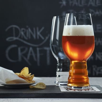 Zwei SPIEGELAU Craft Beer Gläser für IPA auf einem Schiefertablett. Eines davon ist mit IPA-Bier gefüllt, das Glas dahinter ist leer. Dahinter steht ein Teller mit Kartoffelchips und eine Tafel mit einer Bierkarte darauf.<br/>