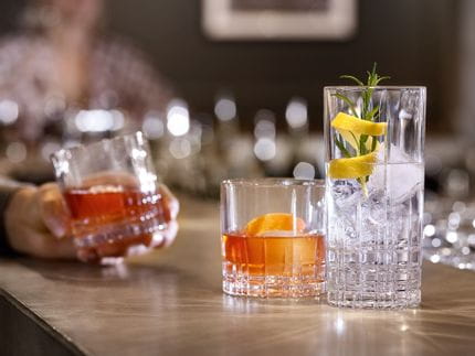 A bar counter with two SPIEGELAU Perfect Serve Collection glasses. The SOF glass is filled with an orange colored drink and an orange zest, the longdrink glass is filled with clear cocktail drink, ice cubes, rosemary and a lemon zest. In the background a hand, holding another filled SOF glass.<br/>