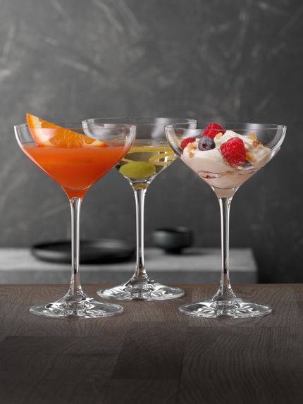 SPIEGELAU Dessert- und Champagnerschalen auf einem Holztisch, jede mit einem anderen Cocktailgetränk gefüllt und eine mit einem Dessert mit Beeren darauf.<br/>