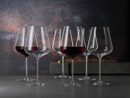 Six SPIEGELAU Definition Bordeaux glasses, two of them are filled with red wine, stand on a table.<br/>