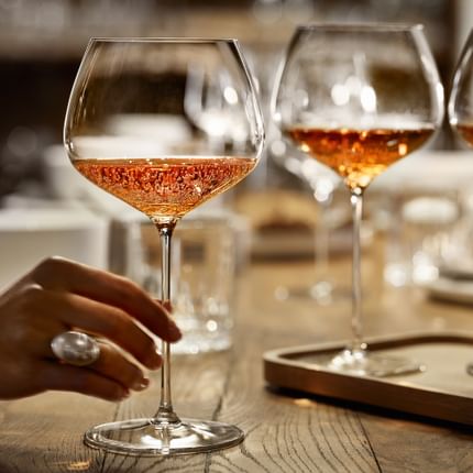 A hand holding the SPIEGELAU Burgundy glass of the series Willsberger Anniversary, filled with rosé colored sparkling wine. Further back on the wooden table, there are more glasses.<br/>