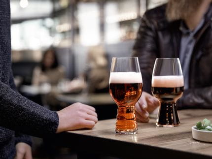 Die SPIEGELAU Craft Beer Gläser IPA Glas gefüllt mit IPA Bier und das Stout Glas gefüllt mit Stout Bier stehen auf einem Holztisch. Dahinter eine Hand, die nach dem Stout-Glas greift.<br/>