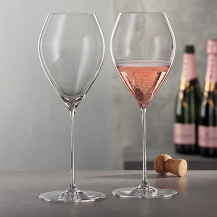 Two SPIEGELAU Spumante glasses standing on a table with a cork in the background. One of the glasses is filled with a pink sparkling wine.<br/>