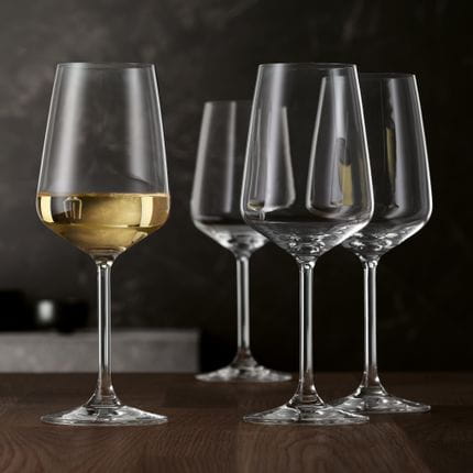 Four SPIEGELAU Style white wine glasses on a wooden table. One glass is filled with white wine.<br/>