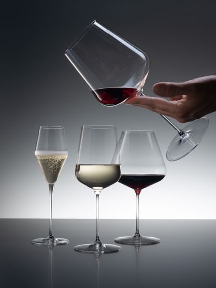Three SPIEGELAU Definition glasses of which the Champagne glass is filled with sparkling wine, the Universal glass with white wine and the Burgundy glass with red wine. Coming from the top right corner, a hand is holding the Bordeaux glass filled with a little red wine.<br/>