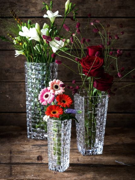Tre vasi di cristallo NACHTMANN Bossa Nova di diverse dimensioni, ciascuno riempito con un diverso bouquet di fiori, su una credenza in legno.<br/>