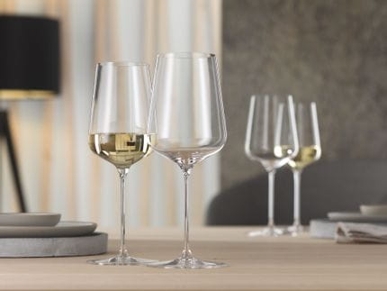 Deux verres de définition SPIEGELAU sont posés sur une table en bois avec deux autres verres de définition en arrière-plan. L'un des verres est rempli de vin blanc, au premier plan et à l'arrière-plan.<br/>