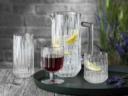 The NACHTMANN Jules crystal glass series on a round serving tray. The pitcher and the tumbler are filled with water, ice cubes and lemon slices, to their right is lavender. The goblet is filled with red wine. The longdrink glass in the background is empty.<br/>