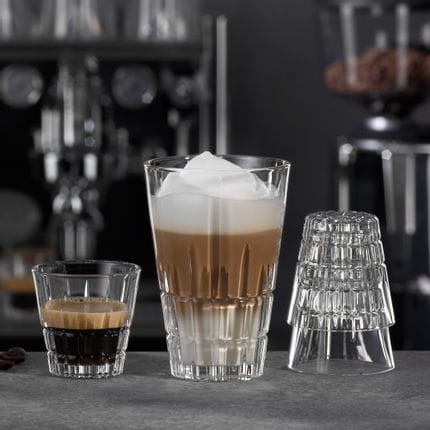 Le verre à espresso de la collection SPIEGELAU Perfect Serve rempli d'espresso et le verre Highball rempli de Latte Macchiato sur un comptoir de bar décoré de grains de café et d'un tamper. En arrière-plan, une machine à café et un moulin.<br/>