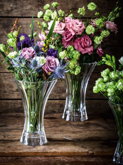 The crystal vases NACHTMANN Saphir, filled with colorful flower bouquets in red, green, violet and blue on a wooden sideboard.<br/>