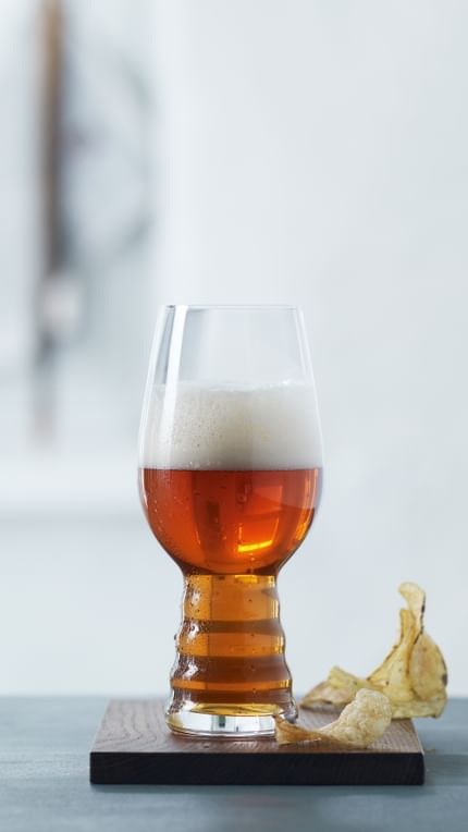 Un gruppo di bicchieri da birra artigianale SPIEGELAU per IPA su un tavolo. Uno è appoggiato su un sottobicchiere di legno, riempito di birra IPA. Accanto a questo bicchiere ci sono delle patatine.<br/>