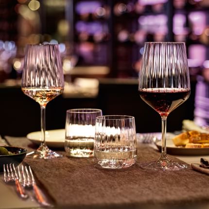 La série SPIEGELAU Lifestyle sur une table de restaurant dans une atmosphère chaleureuse. La lumière violette brille à travers la structure ondulée du verre à vin blanc, du verre à vin rouge, des gobelets à eau et de la carafe.<br/>