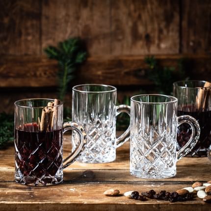 Le verre à thé Noblesse de NACHTMANN contenant du vin chaud et des bâtons de cannelle est posé sur un buffet en bois. À côté se trouvent deux verres à thé Noblesse vides et un verre à thé Noblesse rempli, des amandes, des raisins secs et des branches de pin.<br/>