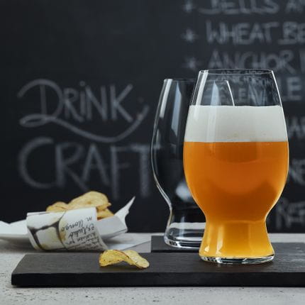 Two SPIEGELAU Craft Beer Glasses for American Wheat Beer and Witbier on slate trays. The glass in the foreground is filled with Wheat Beer, in the background are potato chips and a blackboard with a beer menu on it.<br/>