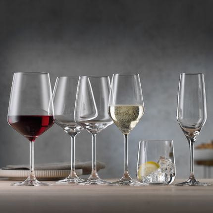 Il bicchiere da vino rosso stile SPIEGELAU pieno accanto al bicchiere da Borgogna vuoto, al bicchiere da vino bianco, al bicchiere da Champagne pieno, al tumbler con acqua, ghiaccio e limone e al flute da Champagne vuoto su un tavolo di legno.<br/>
