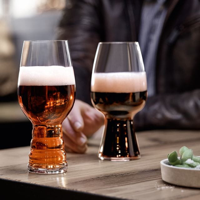 The SPIEGELAU Craft Beer Glasses IPA glass filled with IPA beer and the Stout glass filled with Stout beer standing on a wooden table. Behind them a hand reaching for the Stout glass.<br/>