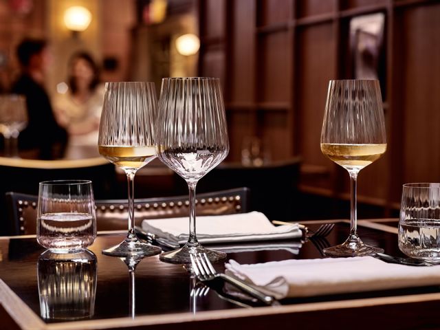 Three SPIEGELAU wine glasses on a laid table. Two of the glasses are filled with white wine, one is empty. To the left of a filled white wine glass is another definition water glass filled with water.<br/>