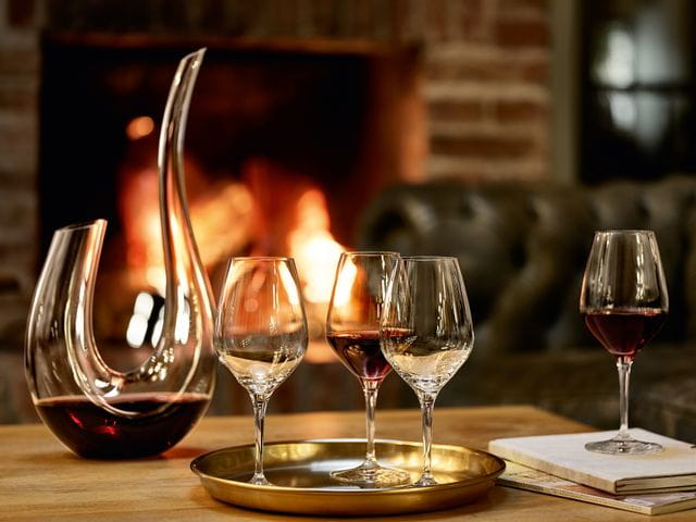A cozy fireplace with three SPIEGELAU Authentis red wine glasses on a serving tray. One glass is filled with red wine. The u shaped hand made decanter SPIEGELAU Novo stands, filled with red wine, on the table as well.<br/>