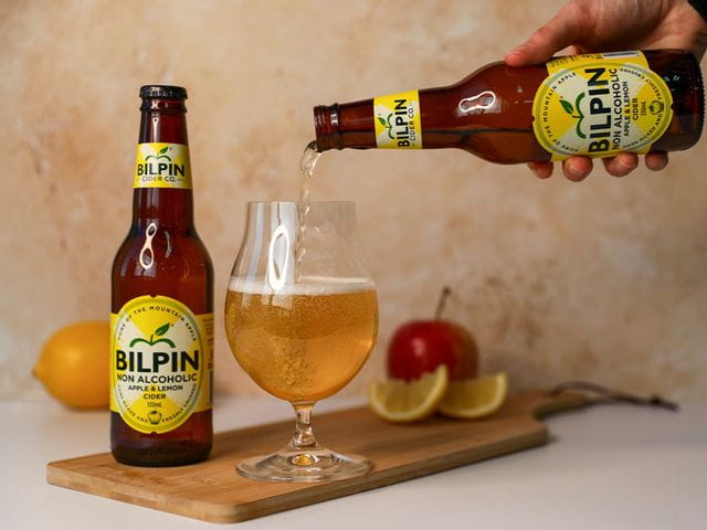 In the middle left of the image is the Spiegelau Beer Classics Tulip glass filled with Bilpin's non-alcoholic cider. To the left of the glass is Bilpin's non-alcoholic cider bottle and above is a hand pouring the bottle into the glass. The bottle and glass sit on a chopping board and in the backdrop is a sliced lemon and apple.