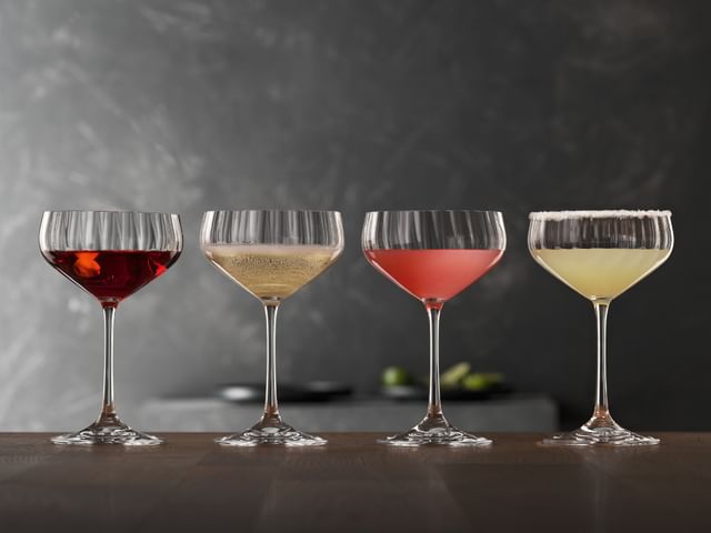 Four SPIEGELAU Lifestyle Coupette glasses on a wooden table. The glasses are filled with different cocktails. From left to right with a Manhattan, a Prosecco, a Cosmopolitan and a Margaritha.<br/>