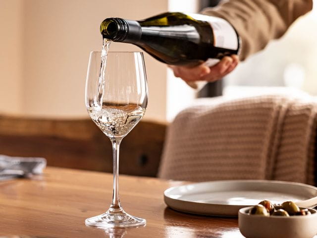 A hand pouring wine into a Spiegelau Vino Grande White Wine glass on a wooden table top.