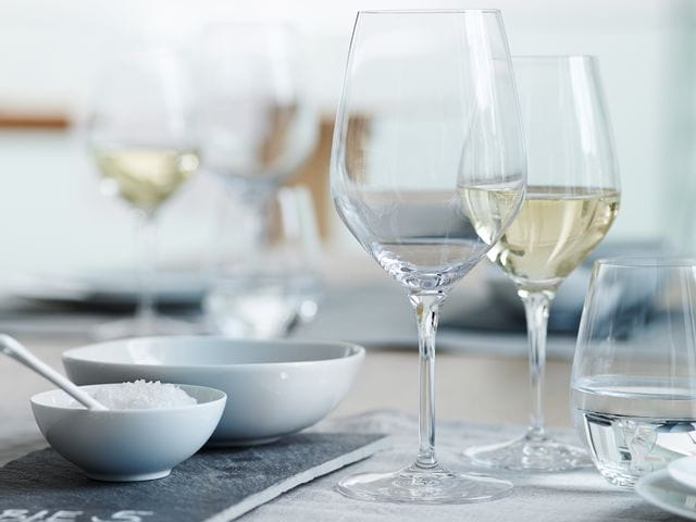 Un bicchiere SPIEGELAU Authentis Bordeaux su un tavolo apparecchiato. Dietro, un bicchiere da vino bianco pieno della linea Authentis e un bicchiere SPIEGELAU Authentis Casual pieno d'acqua, a destra.<br/>