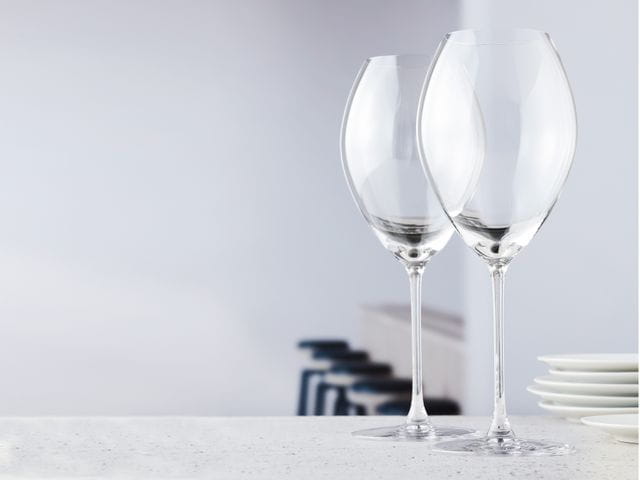 Two empty SPIEGELAU Novo white wine glasses on a sideboard, a row of stools in the background.<br/>