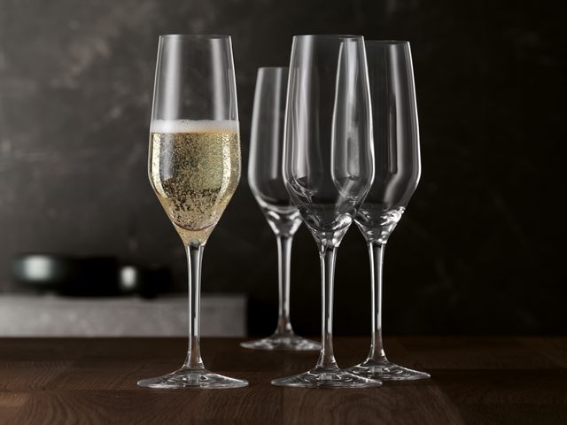 Quattro flauti da champagne in stile SPIEGELAU su un tavolo di legno. Un bicchiere è riempito di spumante.<br/>