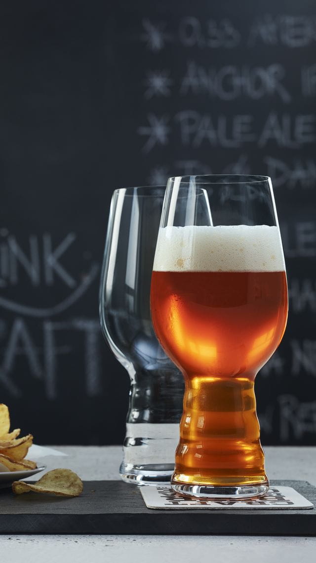 Two SPIEGELAU Craft Beer Glasses for IPA on a slate tray. One of them is filled with IPA beer, the glass behind is empty. Behind them is a plate with potato chips and a blackboard with a beer menu on it.<br/>