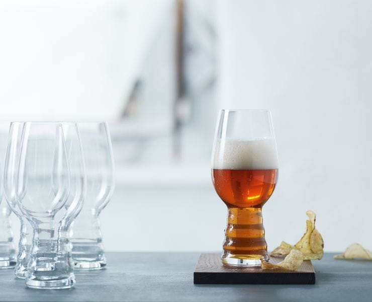 Un gruppo di bicchieri da birra artigianale SPIEGELAU per IPA su un tavolo. Uno è appoggiato su un sottobicchiere di legno, riempito di birra IPA. Accanto a questo bicchiere ci sono delle patatine.<br/>
