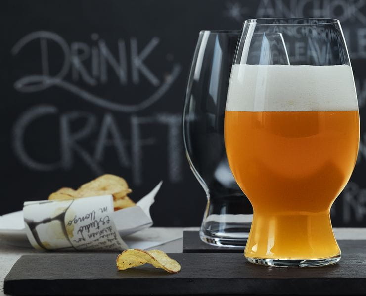 Zwei SPIEGELAU Craft Beer Gläser für American Wheat Beer und Witbier auf Schiefertabletts. Das Glas im Vordergrund ist mit Weißbier gefüllt, im Hintergrund stehen Kartoffelchips und eine Tafel mit einer Bierkarte.<br/>